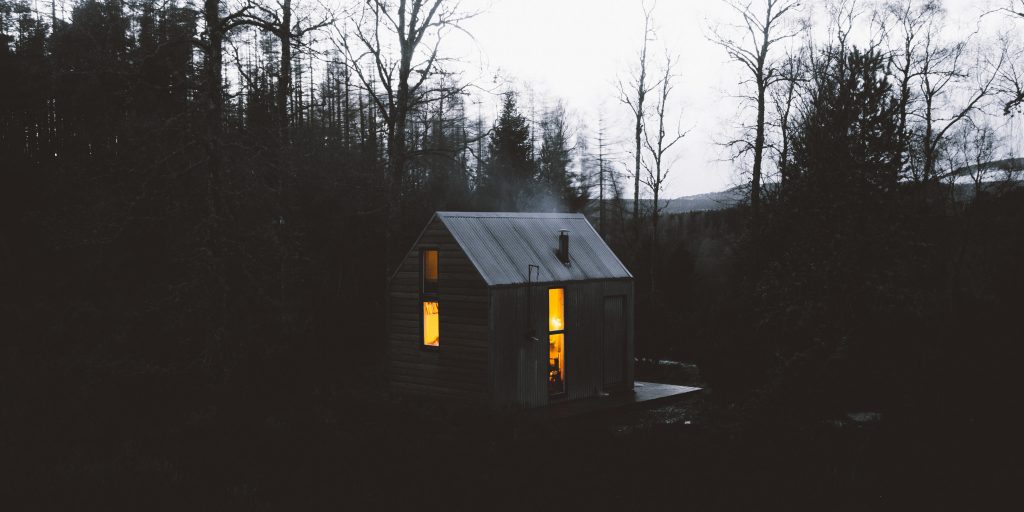 A cabin lit from within at dusk in a remote forest in Scotland.